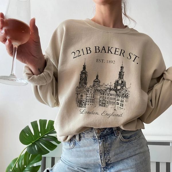 a person with white skin wearing a beige crewneck sweatshirt with black text and design on the front. The text says 221B Baker Street Est 1892 and the design is a London skyline