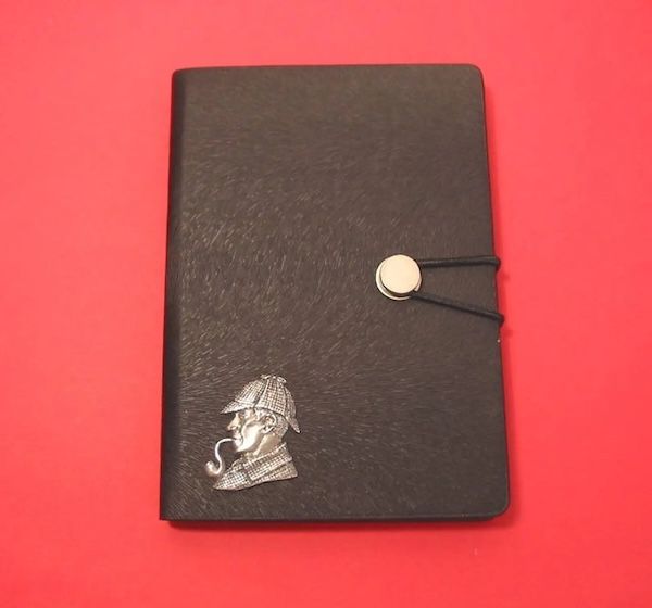 a black notebook resting on a red surface. There is a pewter motif of Sherlock smoking a pipe in profile in the lower left corner
