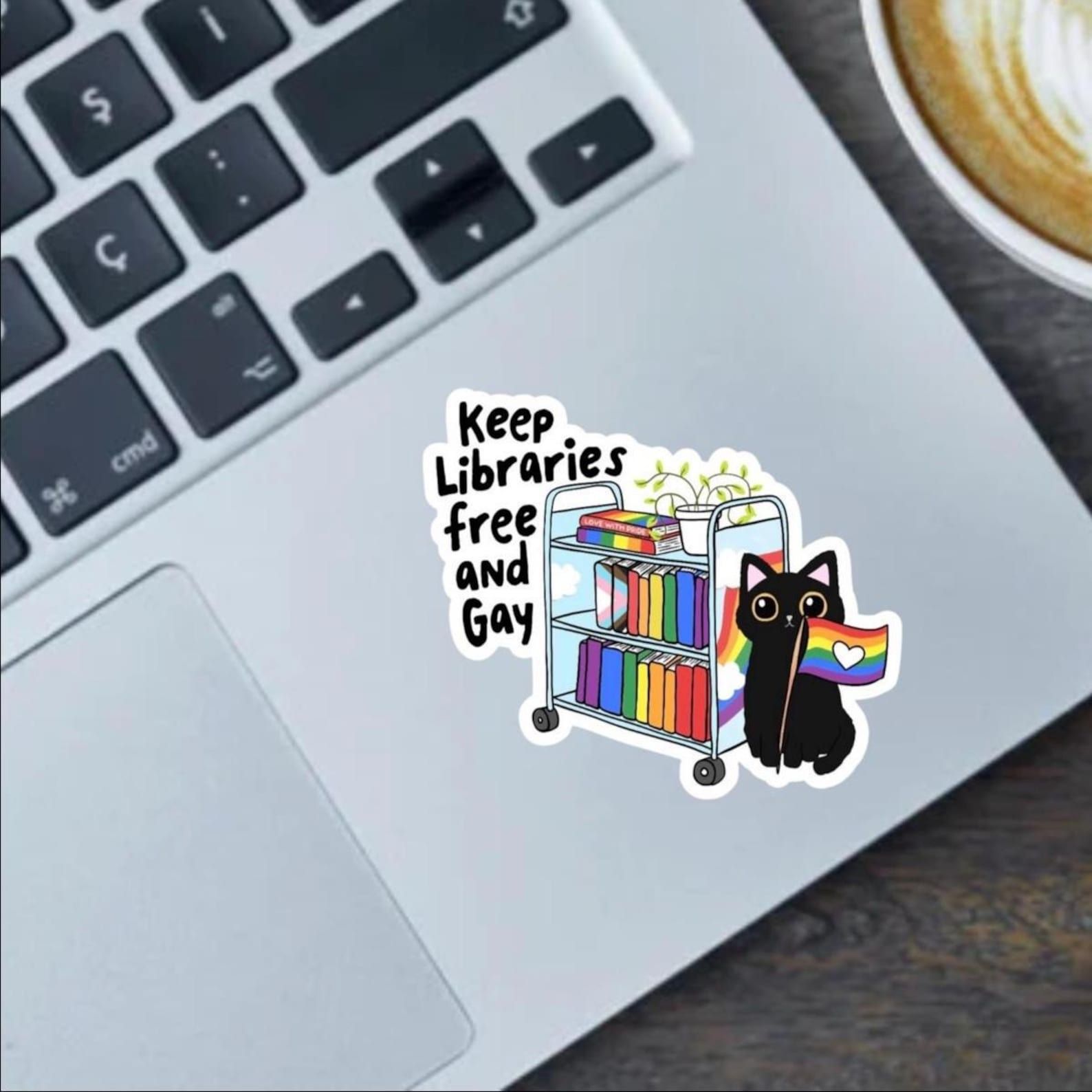 Image of a sticker. It is a bookcart with rainbow book spines on it. There is a black cat with a rainbow pride flag to the cart's left. To the right of the cart are the words "keep libraries free and gay."