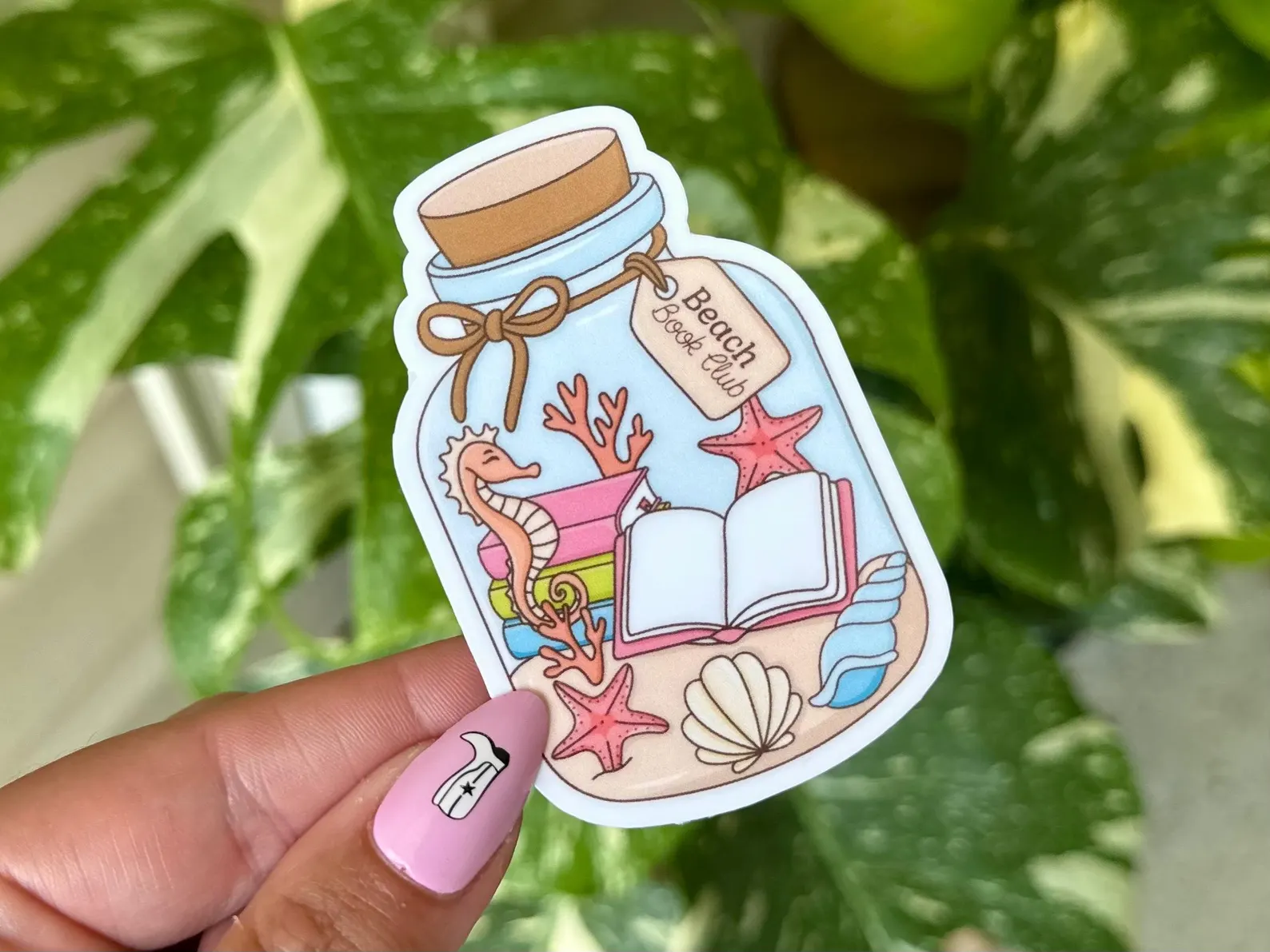Image of a sticker that is a glass bottle. Inside the bottle are books, a seahorse and see shells, and sand. It has a label reading "beach book club."