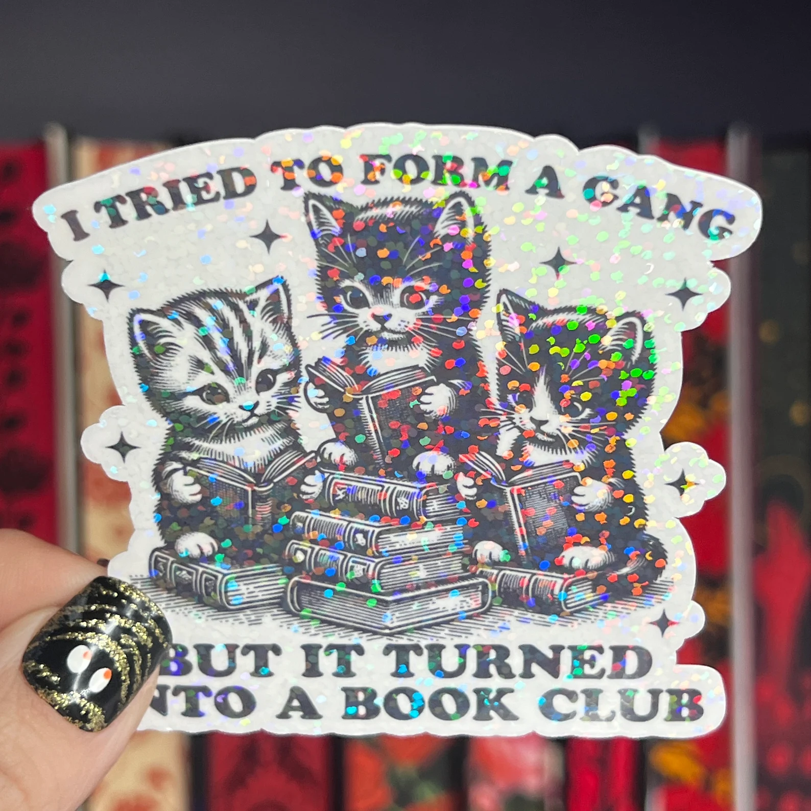 Image of a shiny sticker. It has three cats in the middle reading books. The words around them say "I tried to form a gang but it turned into a book club." 