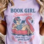 A purple t-shirt that reads 'Book Girl Summer' and 'Dive into a good book' alongside an image of a woman reading while floating on a flamingo tube.