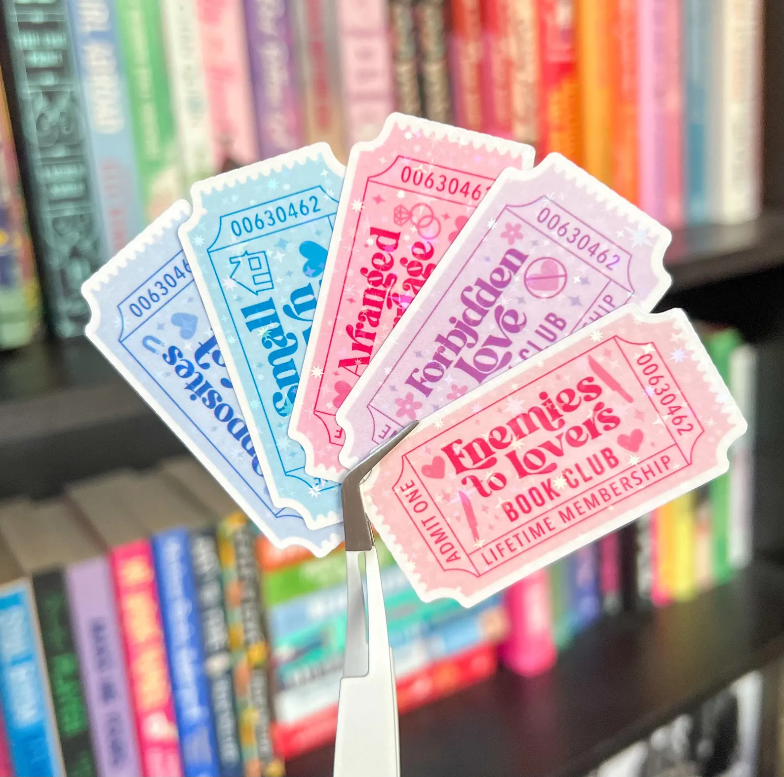 Image of several stickers in the shape of movie tickets. They are in pastel colors and each has a romance trope at the center. For example "Enemies to lovers book club" and "small town romance book club." 