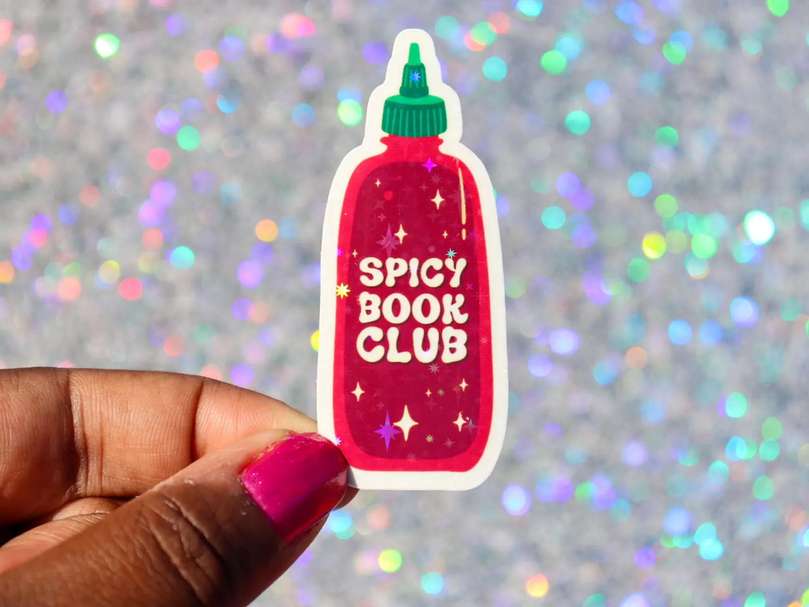 Image of a sticker in the shape of a hot sauce bottle. It's being held by a black hand with pink finger nails. The sticker says "spice book club" on the bottle. 