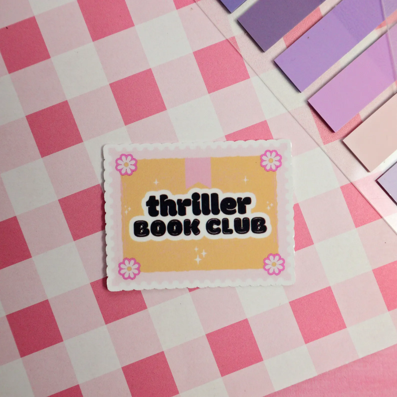 Image of a yellow sticker on a pink checkered background. The sticker has black font that reads "thriller book club."