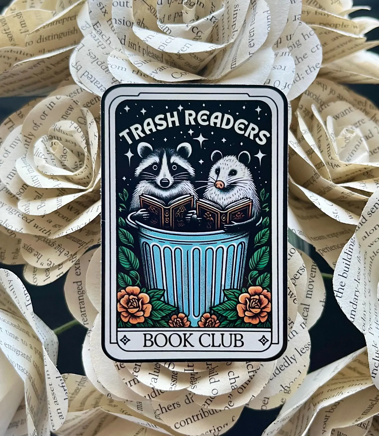 Image of a sticker that looks like a tarot card. There are two raccoons reading books inside a trash can and it has the words "trash readers book club."
