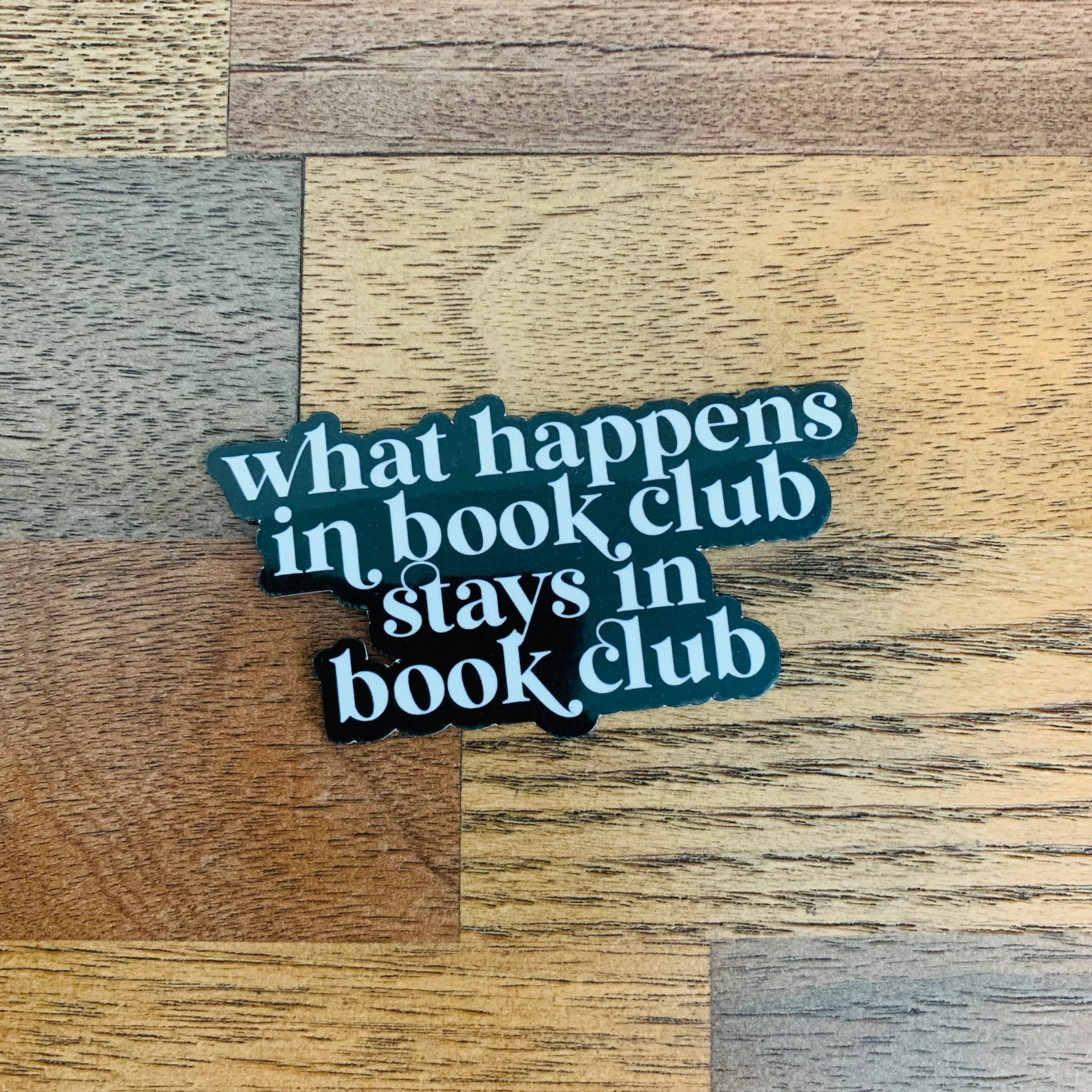 A black sticker with white font on a wood floor. It reads "what happens in book club stays in book club." 