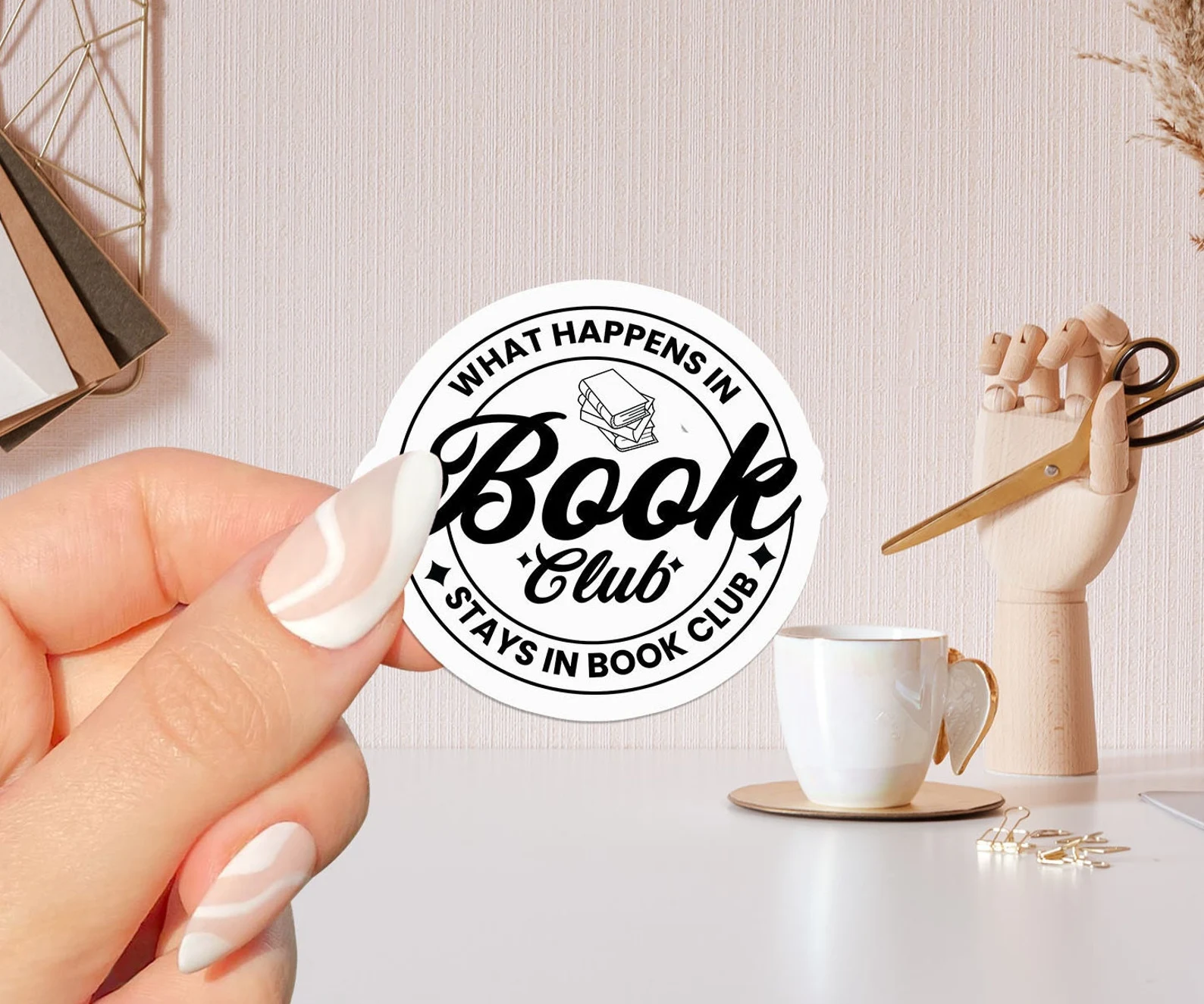 Image of a white hand holding a round white sticker. In black font it reads "what happens in book club stays in book club."