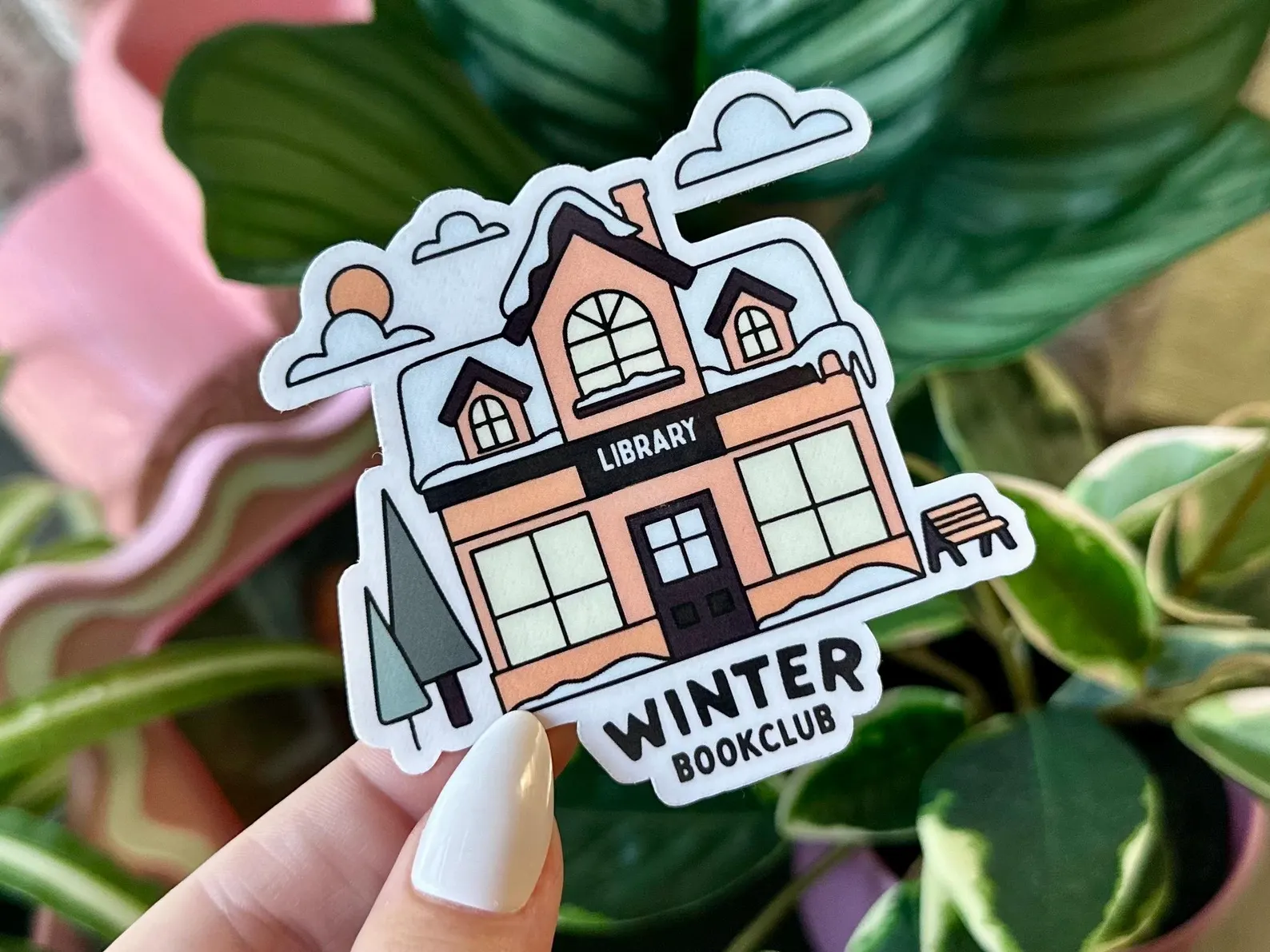 Image of a white hand holding a sticker. It has an image of an orange and black library with snow on the roof. It reads "winter bookclub." 