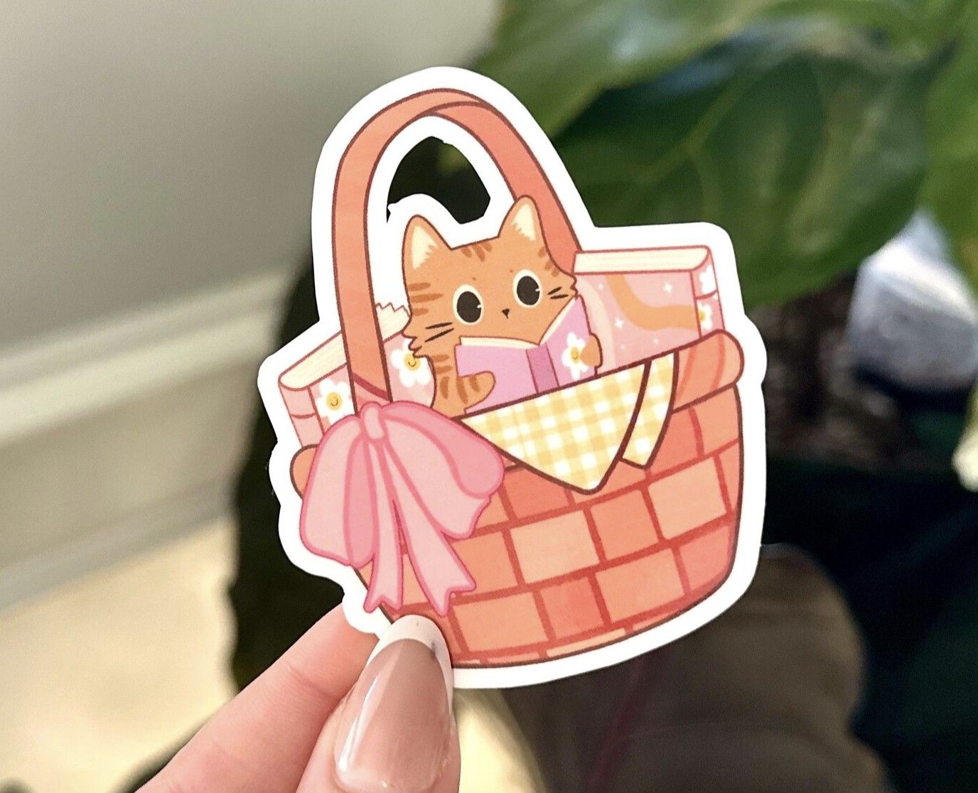 image of a hand holding a vinyl sticker with an illustrated picnic basket and a cat reading a book inside it