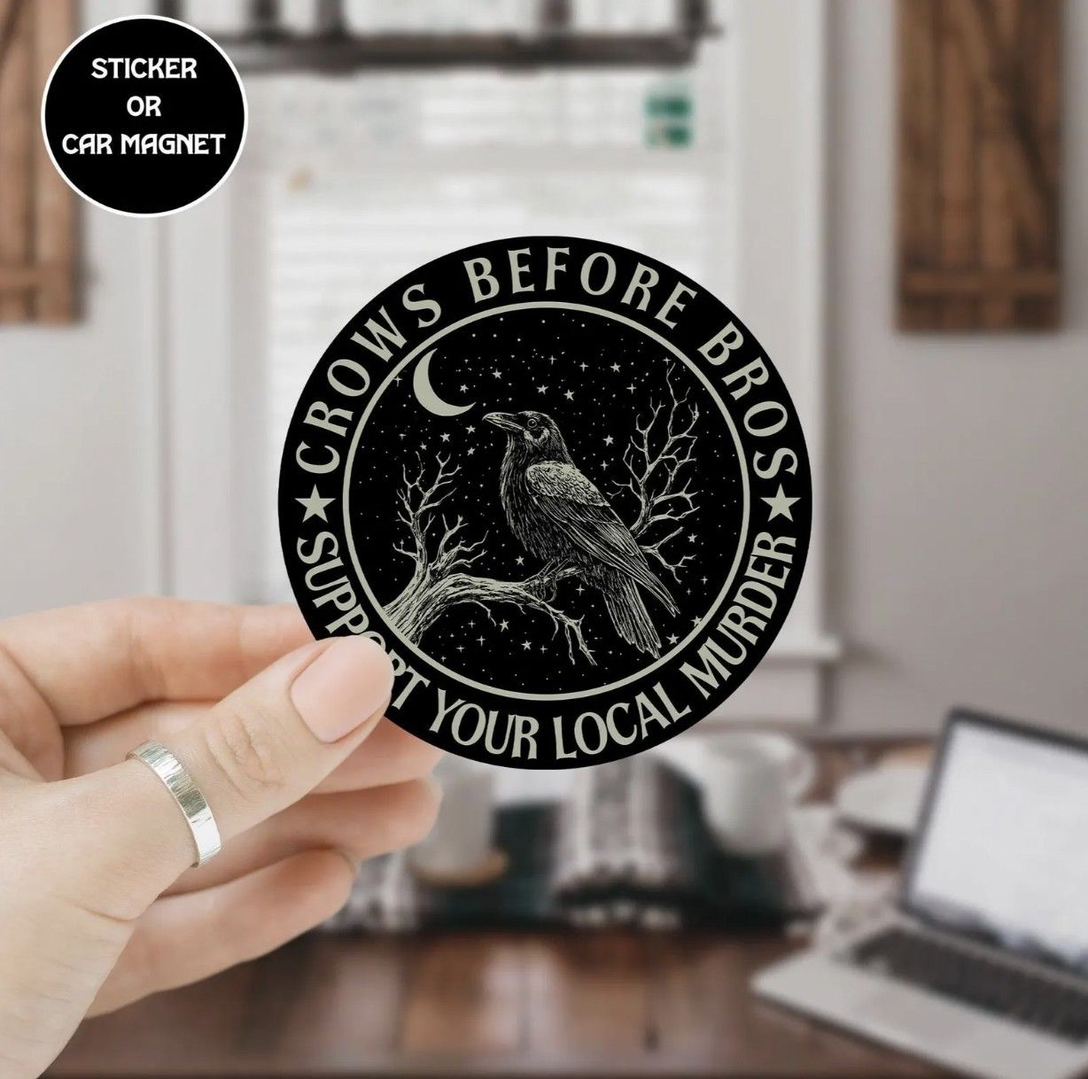 Image of a hand holding a round black sticker with an illustration of a crow and the text "Crows before bros, support your local murder"