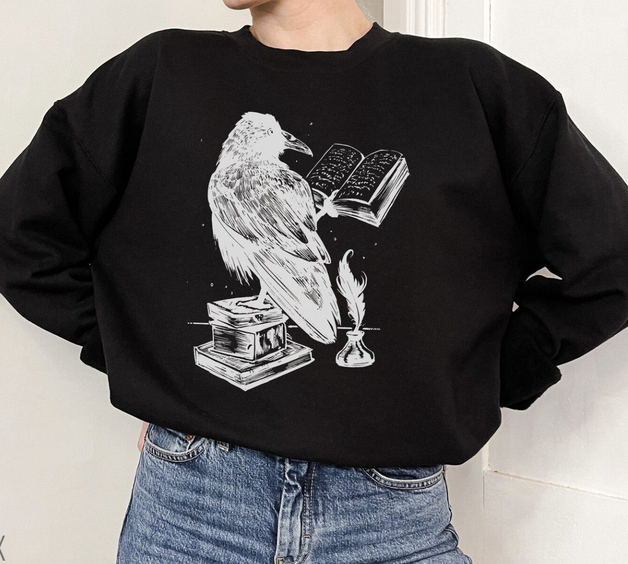 Image of someone modeling a black crewneck sweatshirt with an illustration of a raven reading a book in white