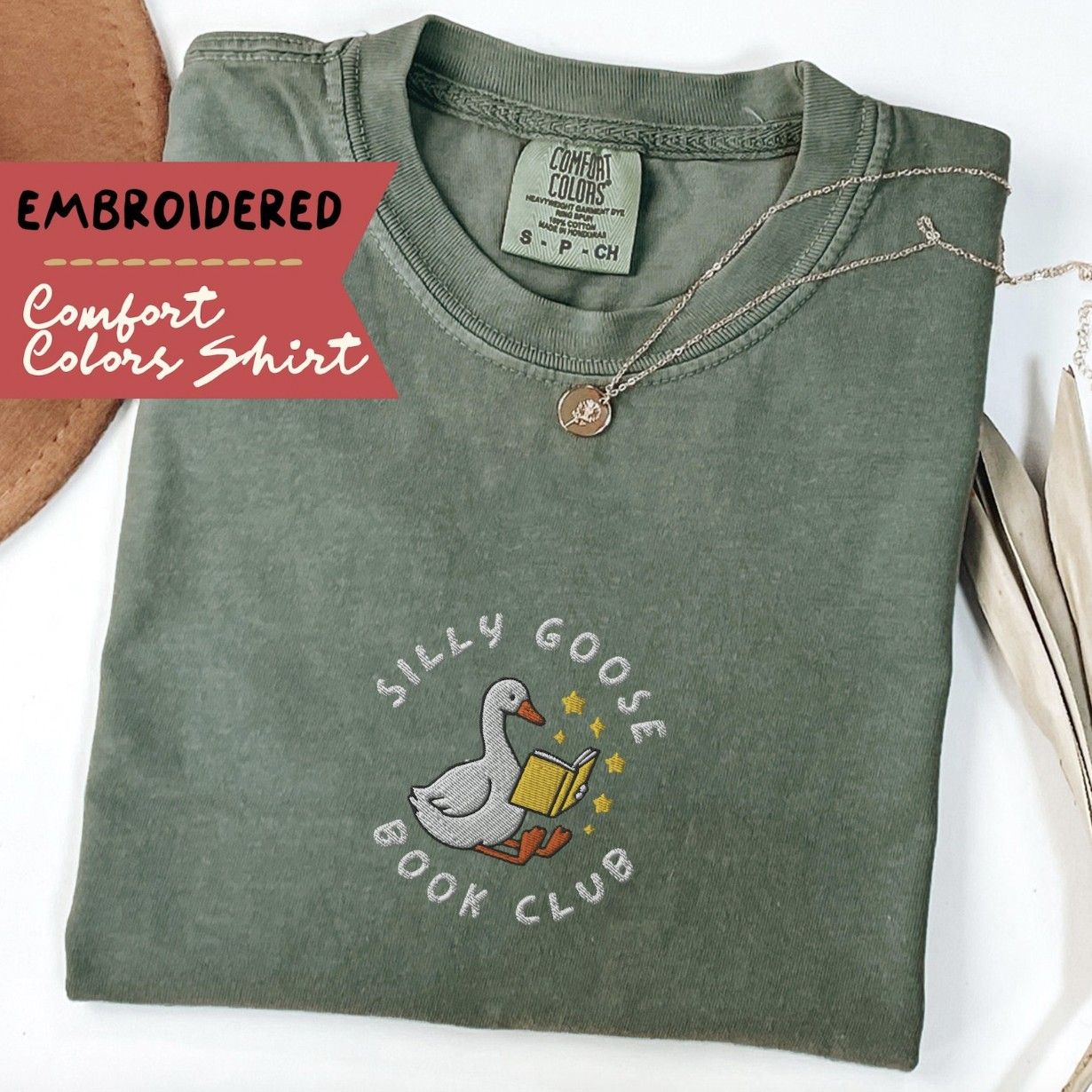 Image of a folded green t-shirt with an embroidered illustration of a goose reading a book and the text "silly goose book club"