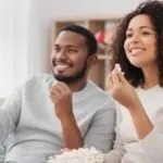 a Black couple watching television on the couch smiling and eating popcorn
