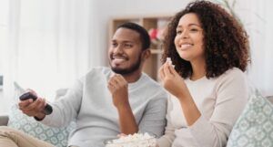 a Black couple watching television on the couch smiling and eating popcorn