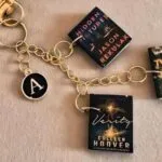 A gold keychain featuring three book titles and a round black initial charm.