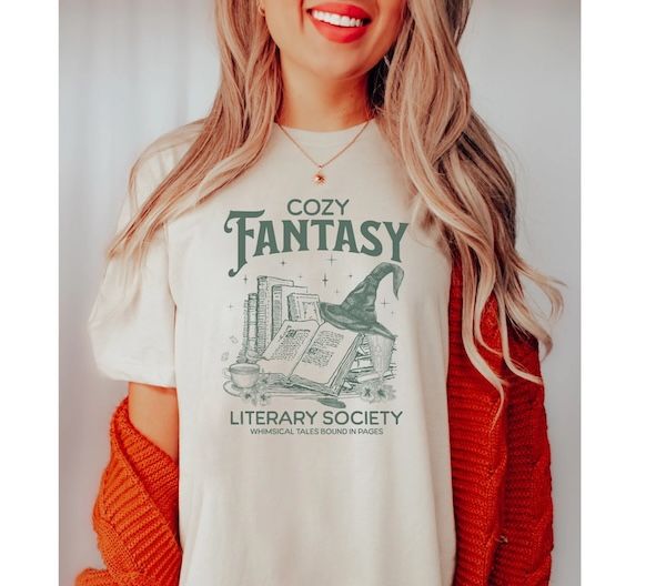Una mujer que usa una camiseta blanca con un texto de comunidad literaria de fantasía relajada verde que rodea un gráfico de algunos libros, una taza de té y un sombrero afilado