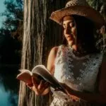 tan skinned woman with hat leaning against a lakeside tree reading