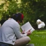 a fair-skinned man reading a book outside on the lawn with swans in the background