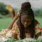 a dark brown skinned woman with braids lays on her stomach reading in the grass