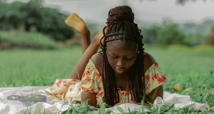 a dark brown skinned woman with braids lays on her stomach reading in the grass