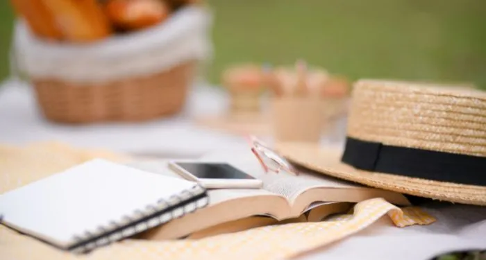 Picnic accessories, a smartphone, a book, and straw picnic hat on a white picnic blanket