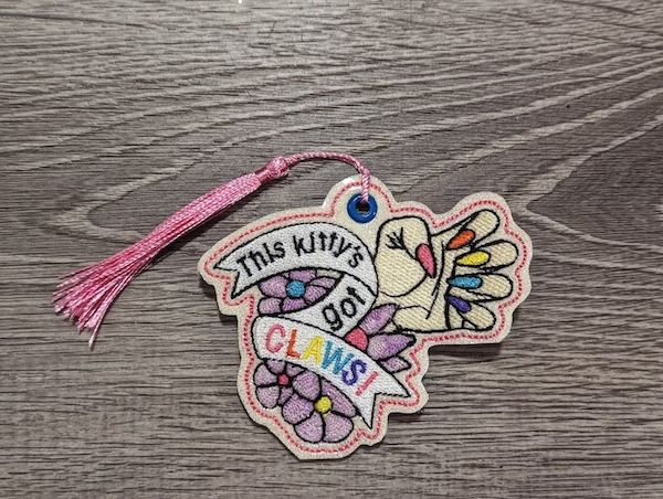 an embroidered glitter vinyl bookmark of a hang with long pointy nails in different colored polish on each nail and text in a banner beneath the hand that reads "this kitty's got claws"