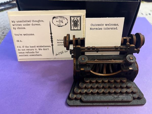 a black mini typewriter positioned next to a postcard. Fed into the typewrite is a small piece of cream-colored paper with black text that reads "outcasts welcome, normies tolerated"