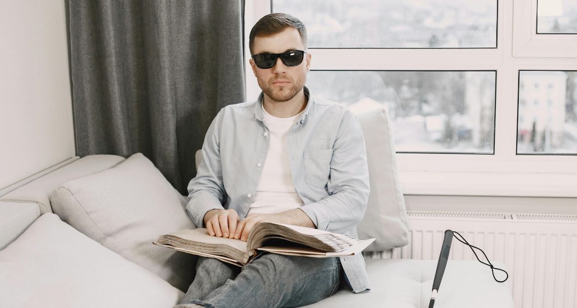 a blind white man sitting on a couch reading a book in braille; his white stick is beside him