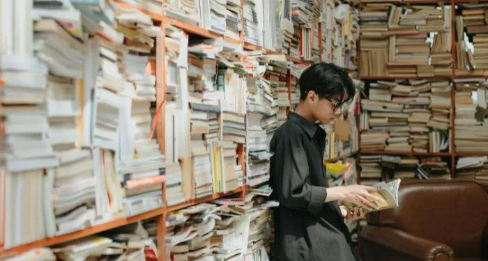 medium tan-skinned Asian man reading a book near shelves of many used books