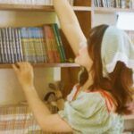a fair-skinned Asian woman reaching for a comic book on a bookshelf