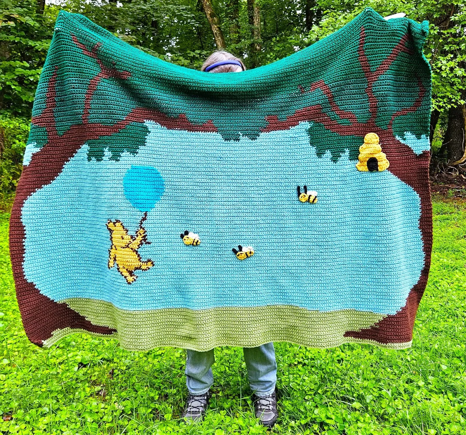 A crochet winnie the pooh blanket held by a person in a yard.