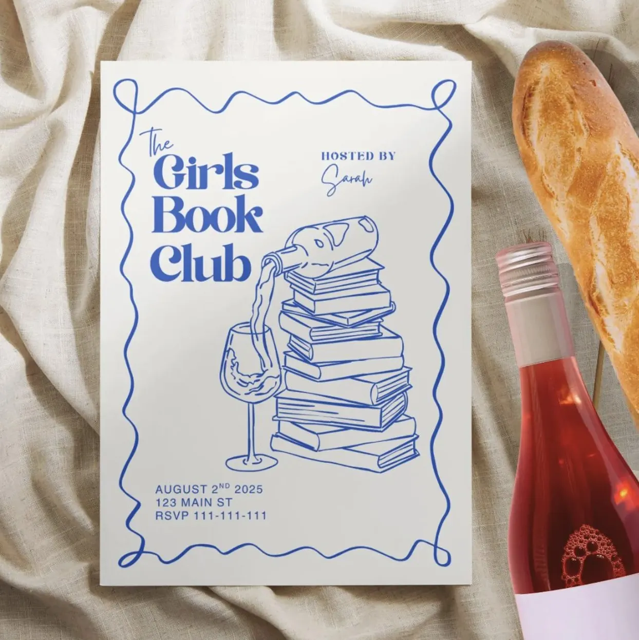 Photo of a white and blue party invitation that says "The Girls Book Club" with an illustration of a stack of books and a wine bottle on top pouring into a wine glass