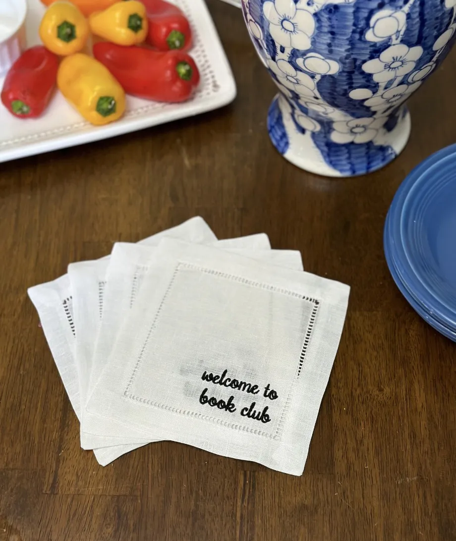 image of four small square linen white napkins with black embroidered cursive text "welcome to book club"