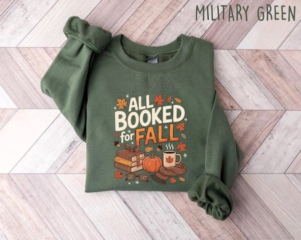 a military green crewneck sweatshirt with text that reads All Book for Fall hovering over graphics of a stack of books, a pumpkin, a steaming cup of coffee, and a scarf. There are leaves in assorted fall colors all over the design