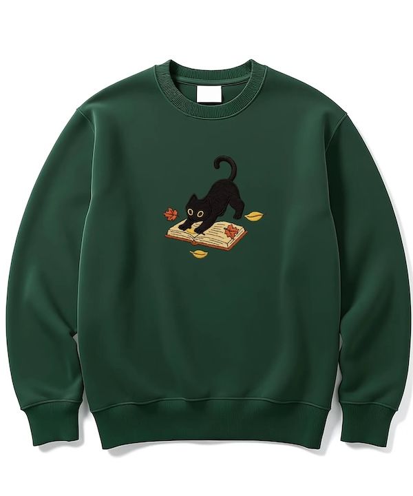 dark green crewneck sweater with an embroidered design of a black cat stretching on top on an open book and red and yellow leaves floating around