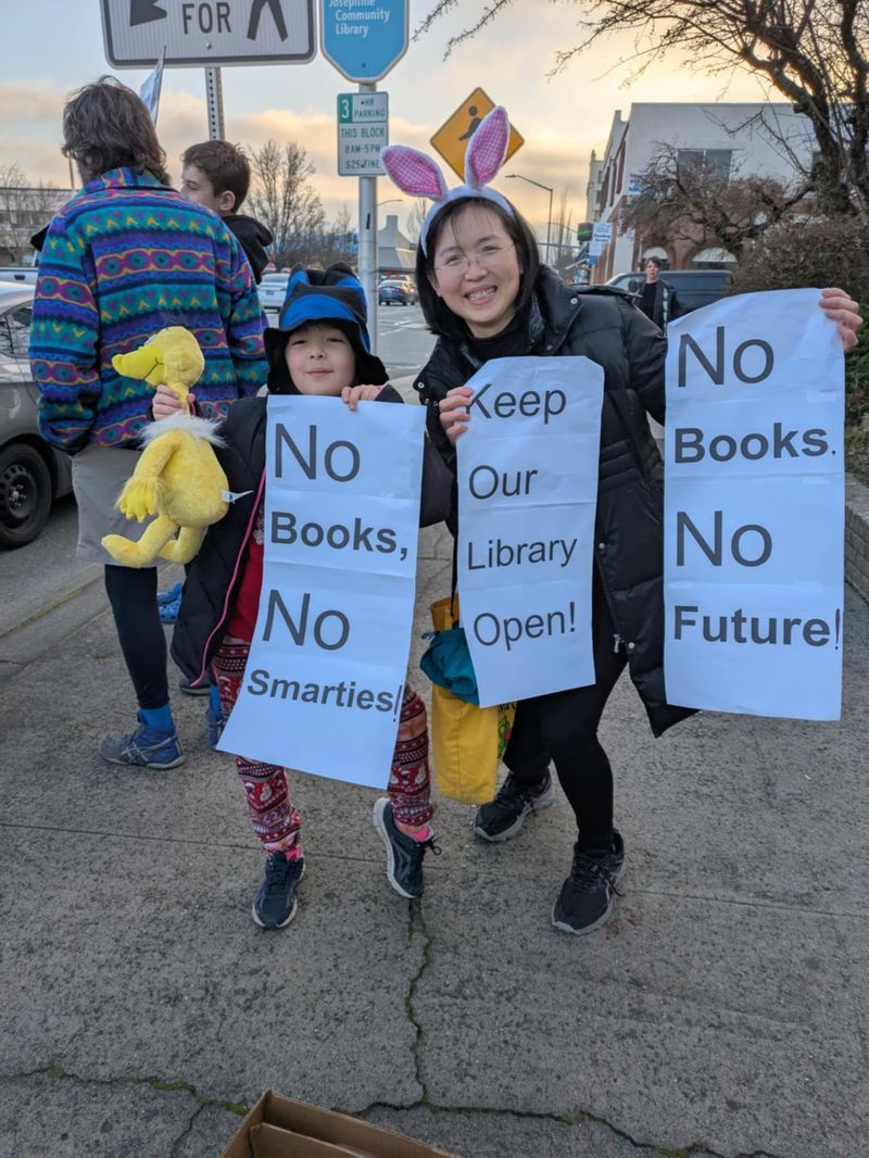Image of an asian woman and child. They are holding signs that read "no books, no smarties," "keep our library open," and "no books, no future." They are at a January 2025 rally for the josephine county library. Image courtesy Image of supporters of josephine county libraries at a january 2025 rally. Image courtesy of Sarah Meyer.