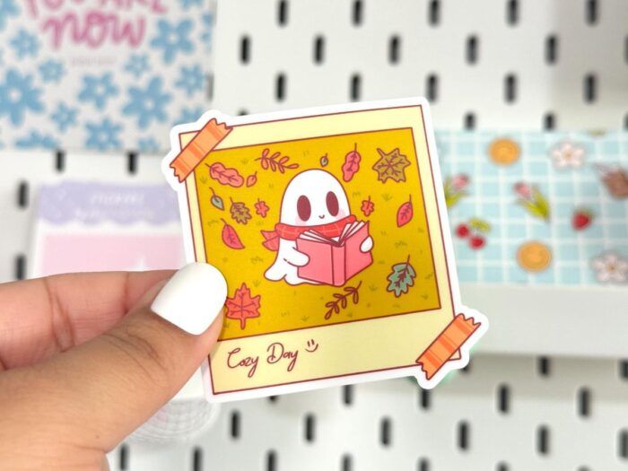 A hand holding a sticker. The sticker is a polaroid with tape on the corners showing a picture of a cartoon ghost reading a book surrounded by fall leaves.
