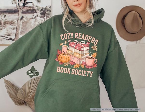 a green hooded sweatshirt with text that reads Cozy Readers Book Society bordering a graphic of a stack of four books with a bow on top. Arranged around the books are a lit pillar candle, a pumpkin, a steaming cup of coffee/tea, and assorted leaves in fall colors