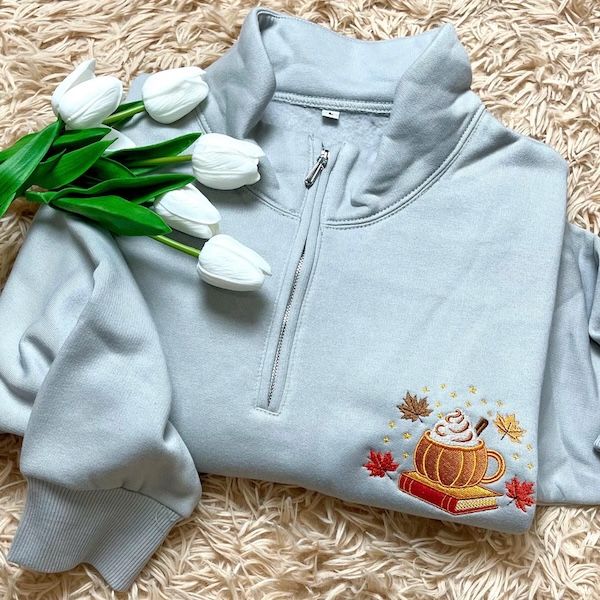 a light blue quarter zip sweatshirt with an embroidered image of a pumpkin-shaped coffee mug on top of a book on the left side. there are four maple leaves positioned around the cup and book