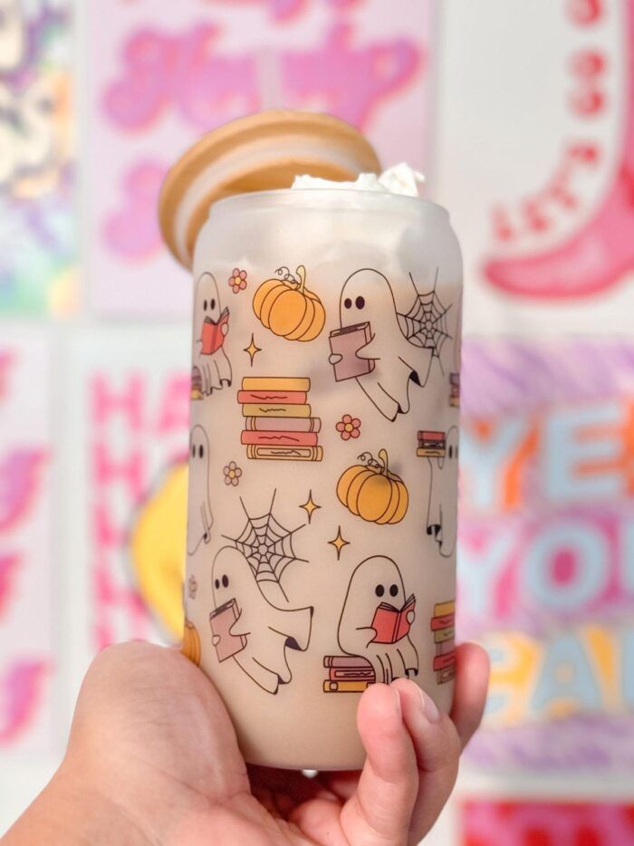 An image of a hand holding a frosted cup decorated with cartoon Halloween decorations like reading ghosts, pumpkins, and spiderwebs