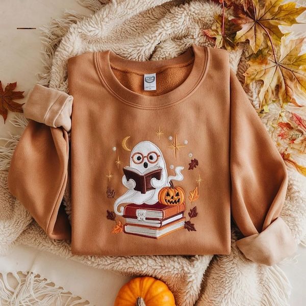 a brown crewneck sweatshirt with an embroidered design of a ghost on the front. the ghost is white and wearing round glasses, and holding a book open while hovering on top of a stack of books. There is pumpkin off to one side and assorted brown and orange leave statrs, and a moon floating around the ghost