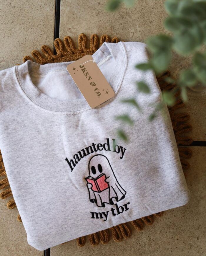 an image of a folded sweatshirt sitting on a table next to a plant. The sweatshirt is embroidered with a cartoon ghost reading and the words "Haunted by my TBR."