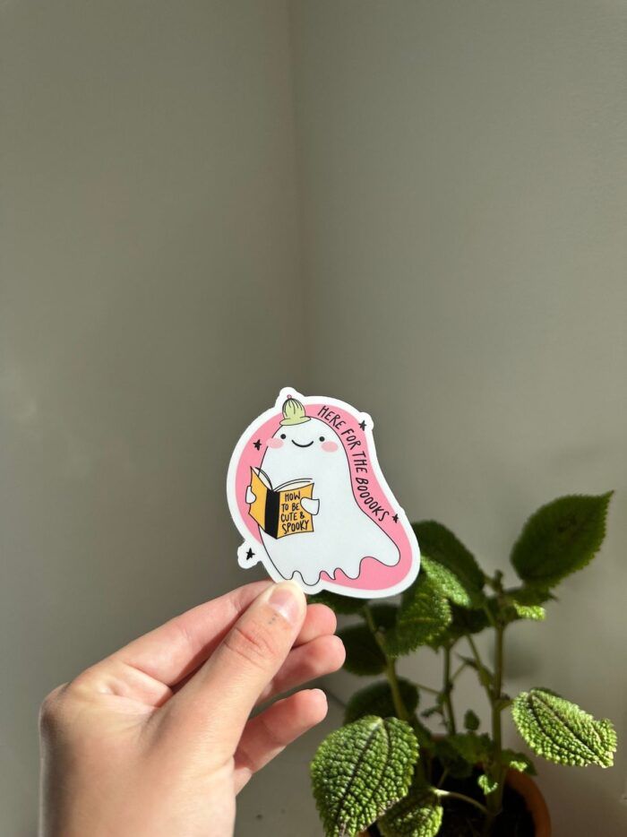 A hand holding a sticker. The sticker is shaped irregularly with a cartoon ghost on a pink background. The ghost is wearing a tiny hat, smiling, and holding a book titled "How To Be Cute and Spooky."