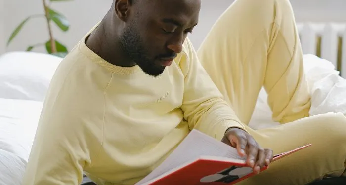 dark brown-skinned Black man in a yellow sweatsuit reading a book in bed