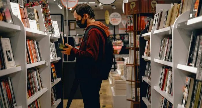 a light-skinned man in a face mask is reading a book in front of a bookshelf