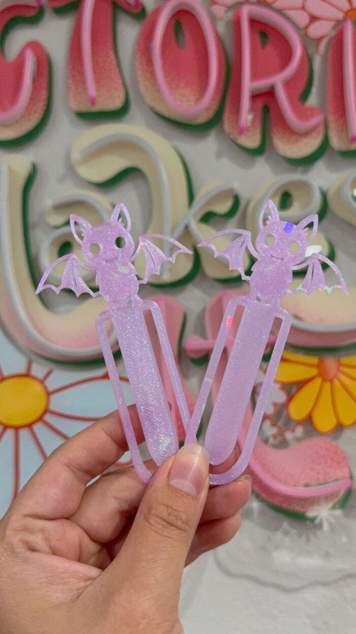 a hand holding two pink plastic bookmarks in the shape of paperclips with a bat design coming off the top of the bookmark
