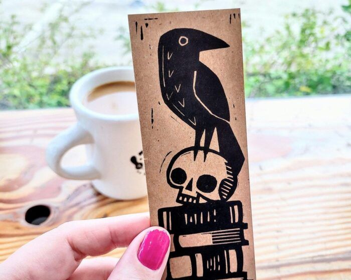 a hand holding a manilla-colored bookmark with a black blockprint image of a skull and a raven on top of a stack of books 