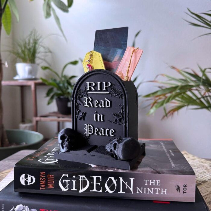 a plastic gravestone-shaped vessel with the words "read in peace." The vessel is holding bookmarks and sitting on top of a copy of Gideon the Ninth