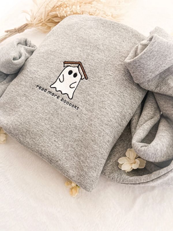heathered grey crewneck sweatshirt with an embroidered ghost on the front. The ghost is white with black trim and there is an open book resting on top of him, and black text beneath him that says "read more booo0ks"